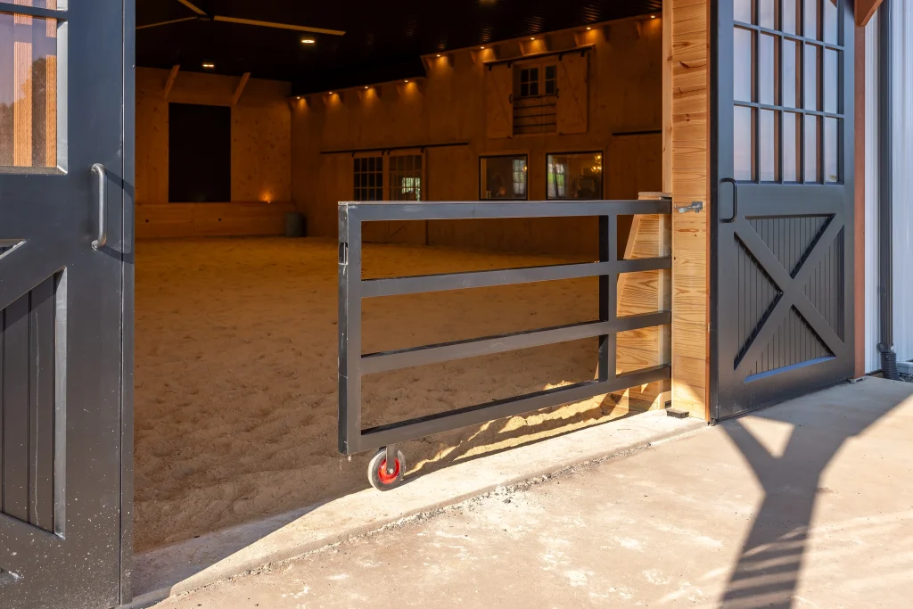 Black steel rolling gate at the entrance to an indoor riding arena with sand footing, wood walls and viewing windows.