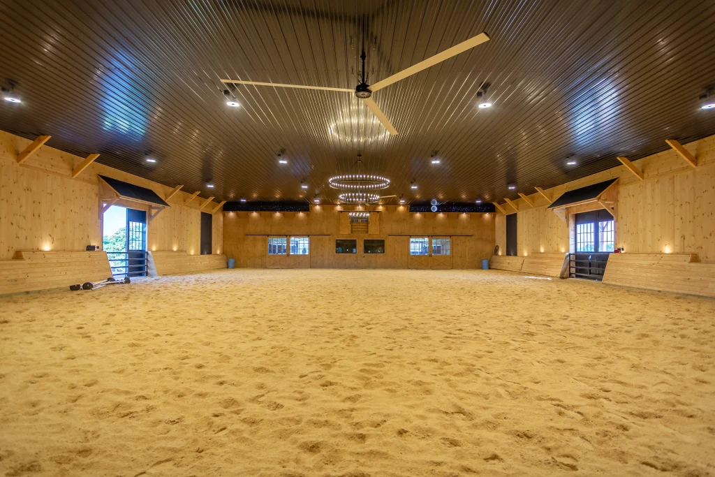 Spacious indoor riding arena with sand footing, light wood-paneled walls, overhead ceiling fans, and hanging chandeliers.