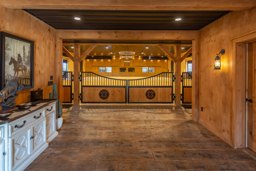 Rustic wooden barn hallway with a white sideboard credenza on the left, looking through decorative wooden and metal gates into an indoor riding arena.